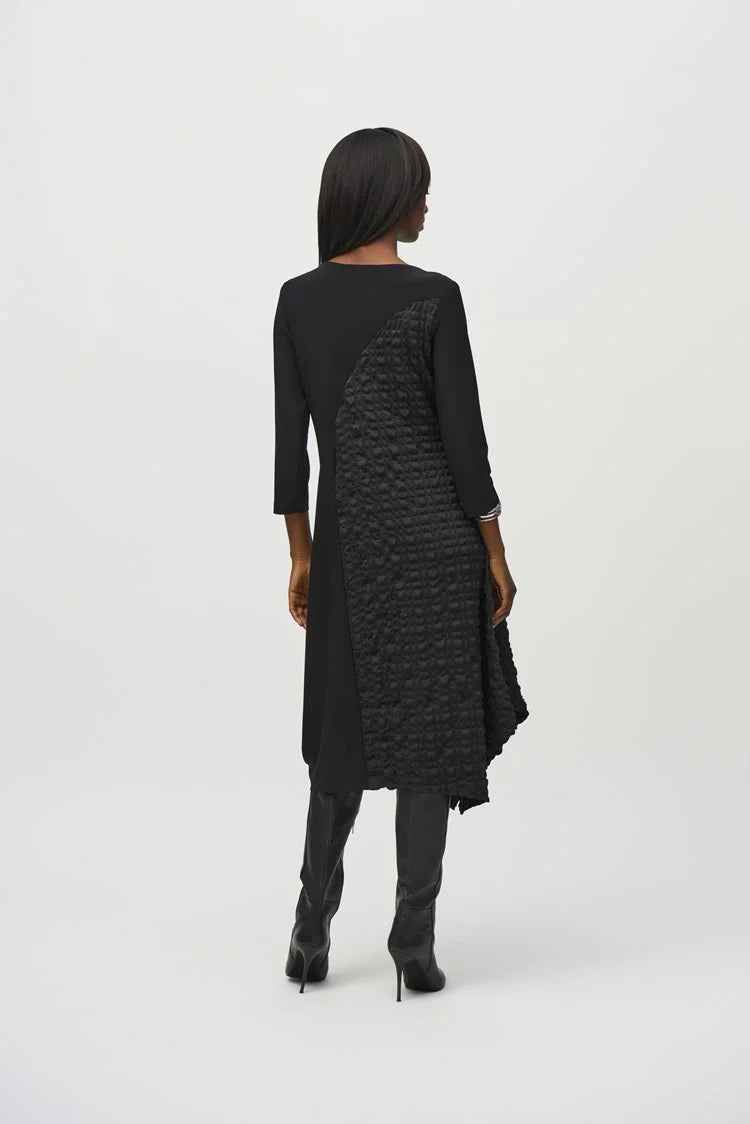 A person with long, straight hair is confidently posing with arms crossed against a plain background. They are wearing the Silky Knit And Jacquard Handkerchief Dress by Joseph Ribkoff, which features an asymmetrical cut and textured pattern on one side. They accessorize with silver bracelets on one wrist.