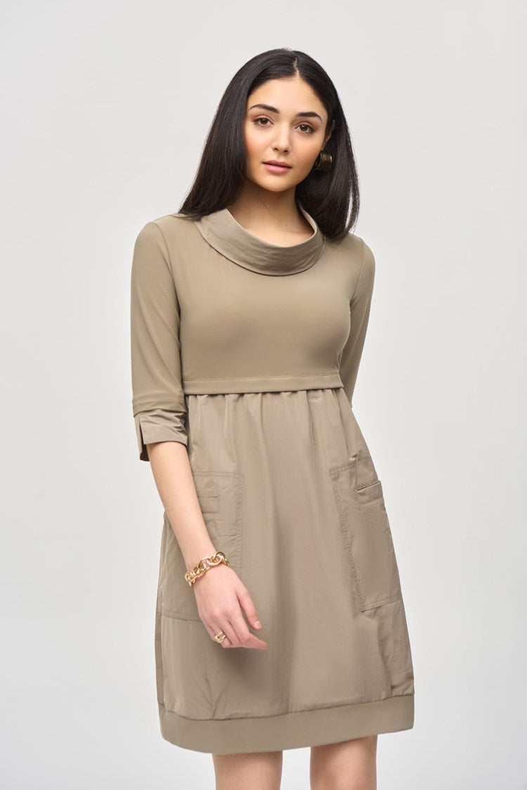 A woman with long dark hair stands against a plain white background, wearing the Cocoon Dress - Seasonal Colors 173444F24 by Joseph Ribkoff. The taupe, long-sleeve, knee-length dress features a cowl neckline and convenient pockets. She pairs it with black knee-high boots and has a neutral expression on her face.