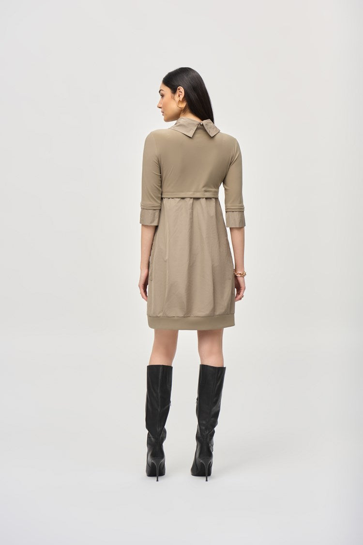 A woman with long dark hair stands against a plain white background, wearing the Cocoon Dress - Seasonal Colors 173444F24 by Joseph Ribkoff. The taupe, long-sleeve, knee-length dress features a cowl neckline and convenient pockets. She pairs it with black knee-high boots and has a neutral expression on her face.