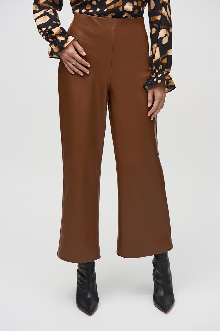 A person is standing indoors, wearing a black and beige patterned top, stylish Joseph Ribkoff Leatherette Wide-Leg Pull-On Pants 244118, and black ankle boots. The backdrop features wood paneling, a leather armchair, and a painting on the wall, creating a warm and sophisticated ambiance.