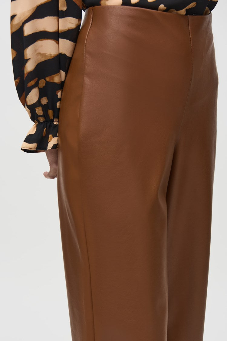 A person is standing indoors, wearing a black and beige patterned top, stylish Joseph Ribkoff Leatherette Wide-Leg Pull-On Pants 244118, and black ankle boots. The backdrop features wood paneling, a leather armchair, and a painting on the wall, creating a warm and sophisticated ambiance.