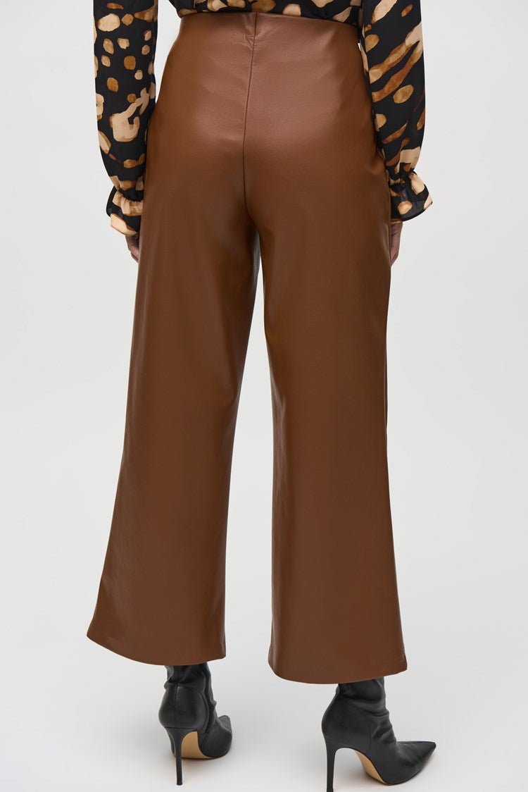 A person is standing indoors, wearing a black and beige patterned top, stylish Joseph Ribkoff Leatherette Wide-Leg Pull-On Pants 244118, and black ankle boots. The backdrop features wood paneling, a leather armchair, and a painting on the wall, creating a warm and sophisticated ambiance.