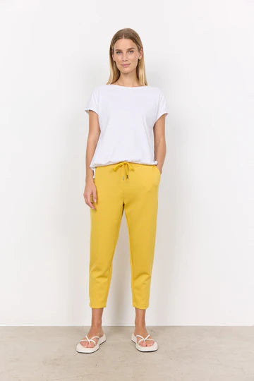 A person with long, blonde hair is wearing a white SC-DERBY 33 T-SHIRT from SoyaConcept, featuring a round neckline. They have paired it with yellow drawstring pants and are standing against a plain, light-colored background. Their expression is neutral as they look directly at the camera.