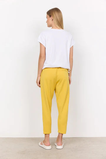 A person with long, blonde hair is wearing a white SC-DERBY 33 T-SHIRT from SoyaConcept, featuring a round neckline. They have paired it with yellow drawstring pants and are standing against a plain, light-colored background. Their expression is neutral as they look directly at the camera.