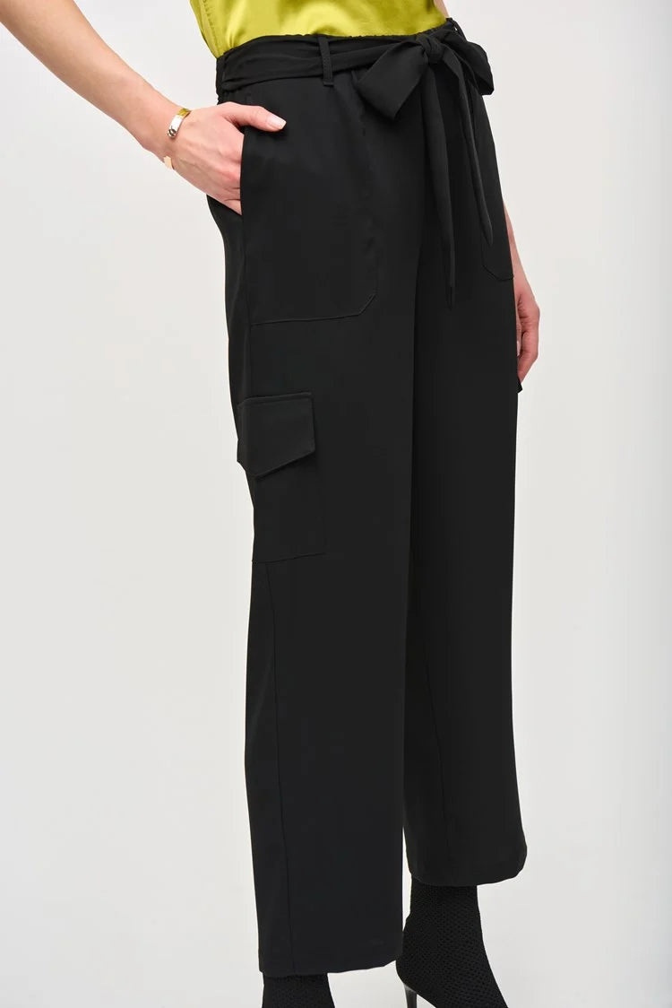 A woman stands against a plain white background, wearing a short-sleeve, lime green blouse paired with the Stretch Woven Pull-On Cargo Pants 243077 by Joseph Ribkoff. The high-waisted, black cropped pants feature a tied belt at the waist. She completes her look with black ankle boots. Her blonde hair is styled in loose waves and she has neutral makeup.