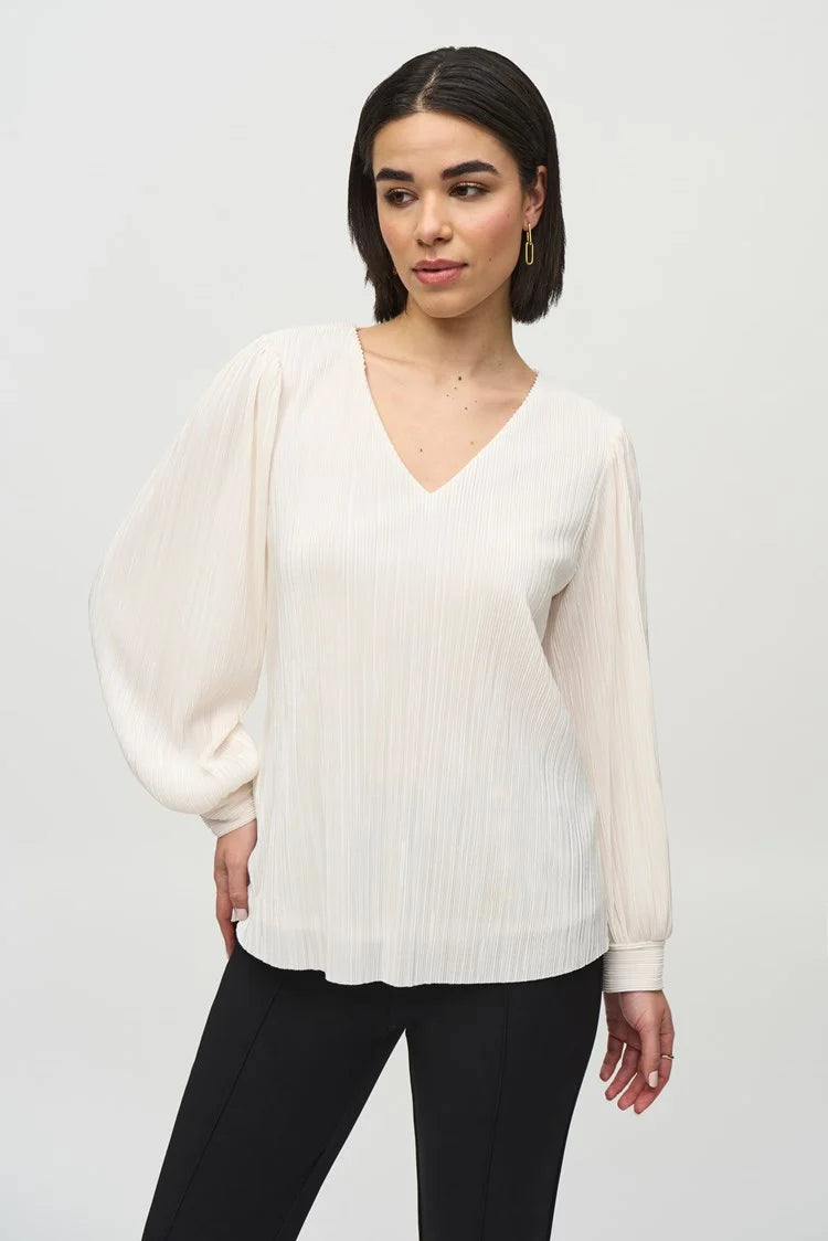 Against a plain background, a person with shoulder-length hair is posing in a long-sleeve, white Pleated Knit Boxy V-Neck Top 244163 by Joseph Ribkoff, which features stylish puff sleeves. They pair the blouse with black trousers and complete the look with black ankle boots. Looking directly at the camera with their arms relaxed by their sides, they exude a casual-chic style.