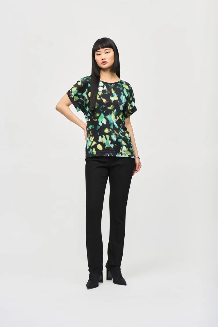 A woman with long, straight black hair and blunt bangs is standing against a plain background. She is wearing the Satin Front Abstract Print Short Sleeve Top (item 243938) by Joseph Ribkoff, which features a knit fabric with a black base adorned with vibrant abstract patterns in green, yellow, and blue hues. She has a neutral expression.
