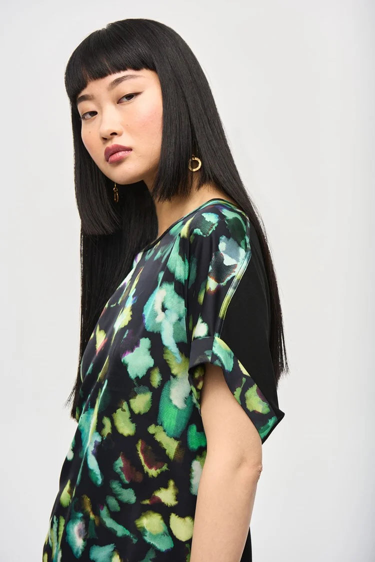 A woman with long, straight black hair and blunt bangs is standing against a plain background. She is wearing the Satin Front Abstract Print Short Sleeve Top (item 243938) by Joseph Ribkoff, which features a knit fabric with a black base adorned with vibrant abstract patterns in green, yellow, and blue hues. She has a neutral expression.