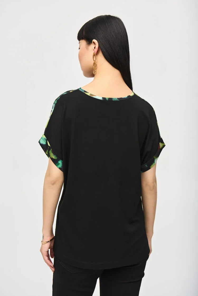 A woman with long, straight black hair and blunt bangs is standing against a plain background. She is wearing the Satin Front Abstract Print Short Sleeve Top (item 243938) by Joseph Ribkoff, which features a knit fabric with a black base adorned with vibrant abstract patterns in green, yellow, and blue hues. She has a neutral expression.