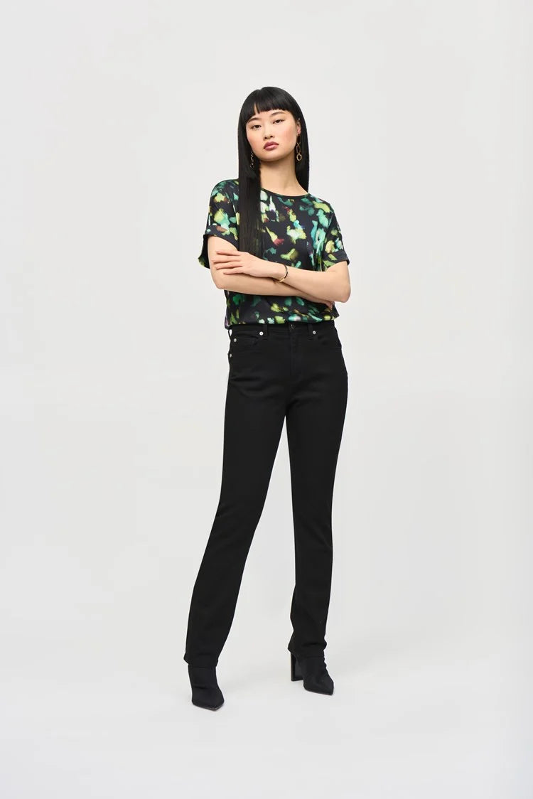 A woman with long, straight black hair and blunt bangs is standing against a plain background. She is wearing the Satin Front Abstract Print Short Sleeve Top (item 243938) by Joseph Ribkoff, which features a knit fabric with a black base adorned with vibrant abstract patterns in green, yellow, and blue hues. She has a neutral expression.