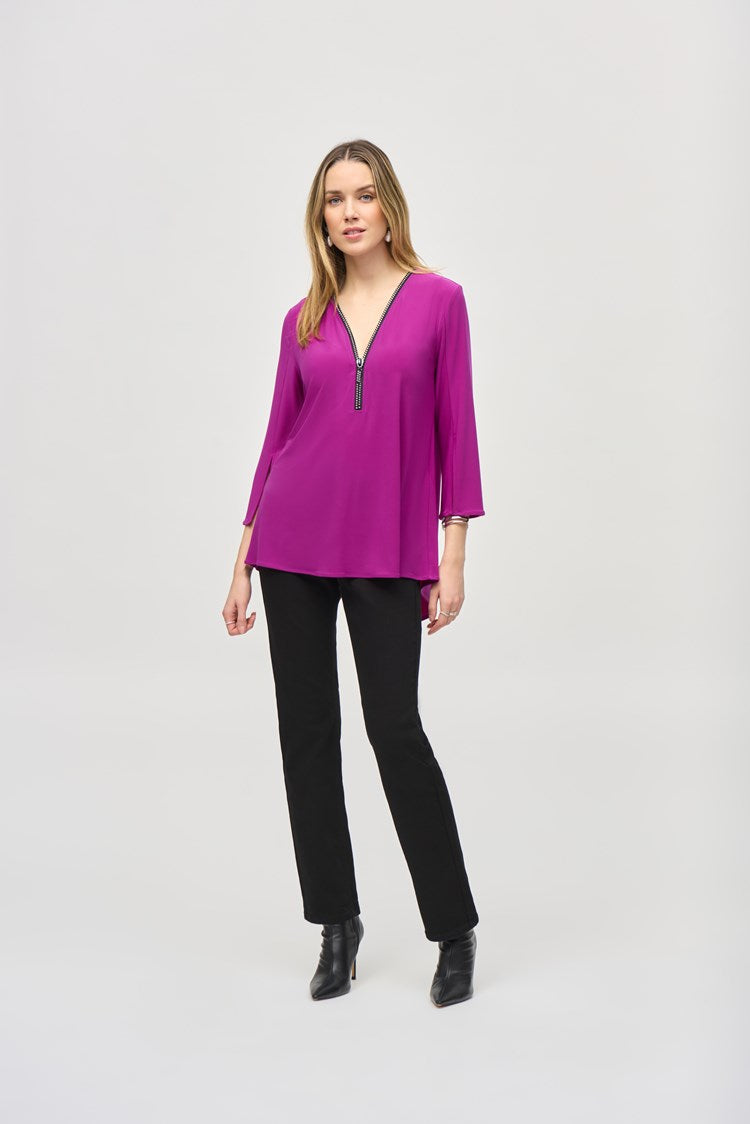 A woman with long hair is standing against a plain background, wearing the Silky Knit Fit and Flare Tunic 243314 by Joseph Ribkoff. The tunic features a long-sleeved, purple V-neck design made of silky knit fabric and adorned with a Rhinestone zipper neckline. She pairs it with black pants and black boots, showcasing a relaxed and confident posture.