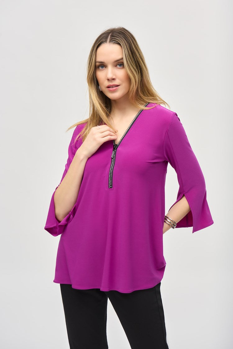 A woman with long hair is standing against a plain background, wearing the Silky Knit Fit and Flare Tunic 243314 by Joseph Ribkoff. The tunic features a long-sleeved, purple V-neck design made of silky knit fabric and adorned with a Rhinestone zipper neckline. She pairs it with black pants and black boots, showcasing a relaxed and confident posture.