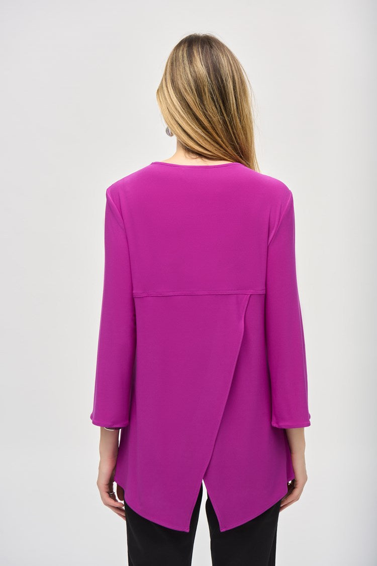A woman with long hair is standing against a plain background, wearing the Silky Knit Fit and Flare Tunic 243314 by Joseph Ribkoff. The tunic features a long-sleeved, purple V-neck design made of silky knit fabric and adorned with a Rhinestone zipper neckline. She pairs it with black pants and black boots, showcasing a relaxed and confident posture.