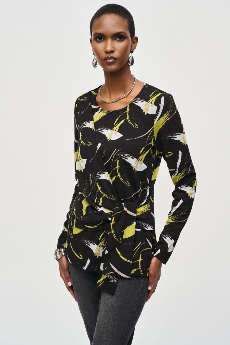 A person stands confidently against a plain white background, wearing a Joseph Ribkoff Sweater Knit Abstract Print Top 243197 featuring a black long-sleeve design with yellow and white abstract patterns and front draping. They pair it with black pants and black heeled boots. Their short hair is styled to highlight the top's scoop neckline, accessorized with earrings, striking a poised pose.