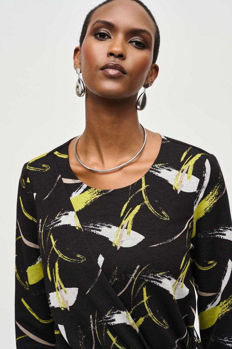 A person stands confidently against a plain white background, wearing a Joseph Ribkoff Sweater Knit Abstract Print Top 243197 featuring a black long-sleeve design with yellow and white abstract patterns and front draping. They pair it with black pants and black heeled boots. Their short hair is styled to highlight the top's scoop neckline, accessorized with earrings, striking a poised pose.