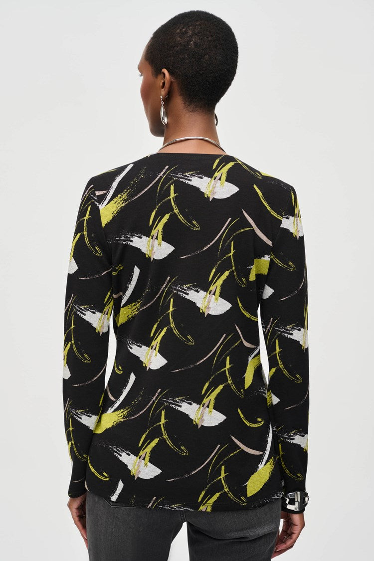 A person stands confidently against a plain white background, wearing a Joseph Ribkoff Sweater Knit Abstract Print Top 243197 featuring a black long-sleeve design with yellow and white abstract patterns and front draping. They pair it with black pants and black heeled boots. Their short hair is styled to highlight the top's scoop neckline, accessorized with earrings, striking a poised pose.