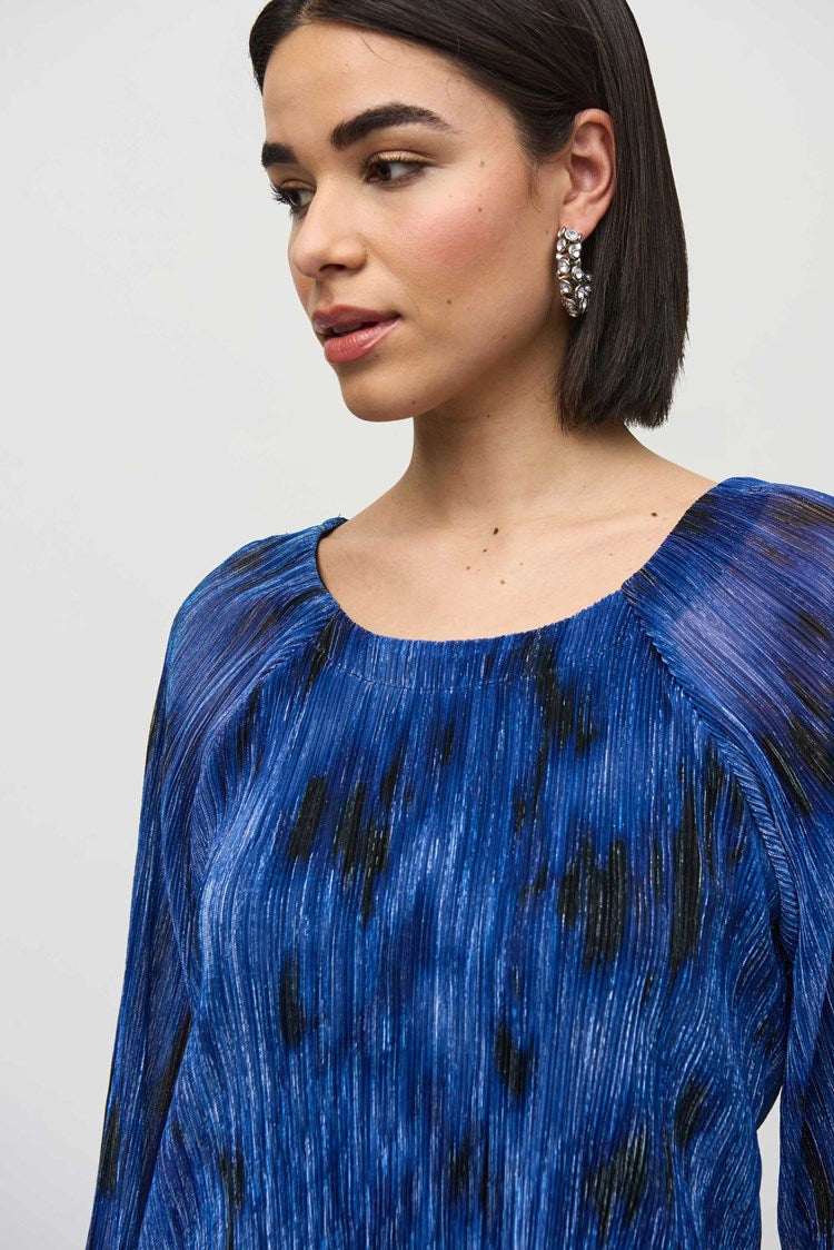 A person with shoulder-length black hair is standing against a plain background, wearing the Joseph Ribkoff Pleated Knit Abstract Print Flared Top 244105, which is blue with long sleeves and features a textured, pleated design. They are also wearing dark pants and are looking slightly to the side with a calm expression.