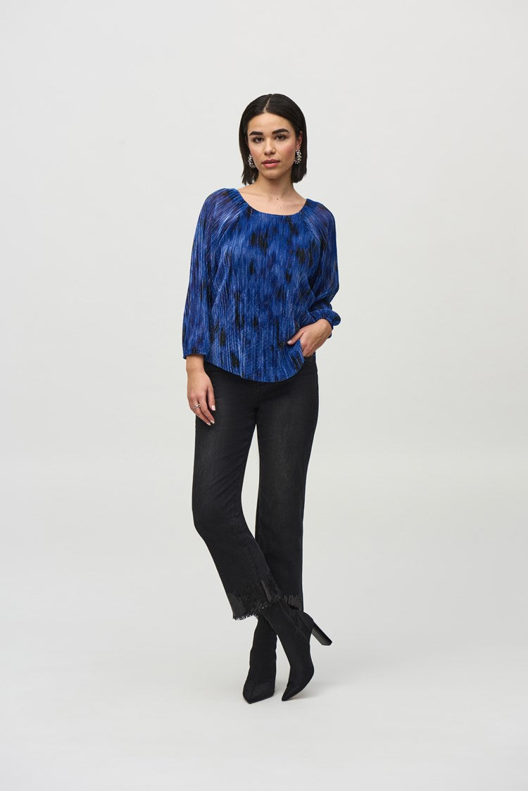 A person with shoulder-length black hair is standing against a plain background, wearing the Joseph Ribkoff Pleated Knit Abstract Print Flared Top 244105, which is blue with long sleeves and features a textured, pleated design. They are also wearing dark pants and are looking slightly to the side with a calm expression.