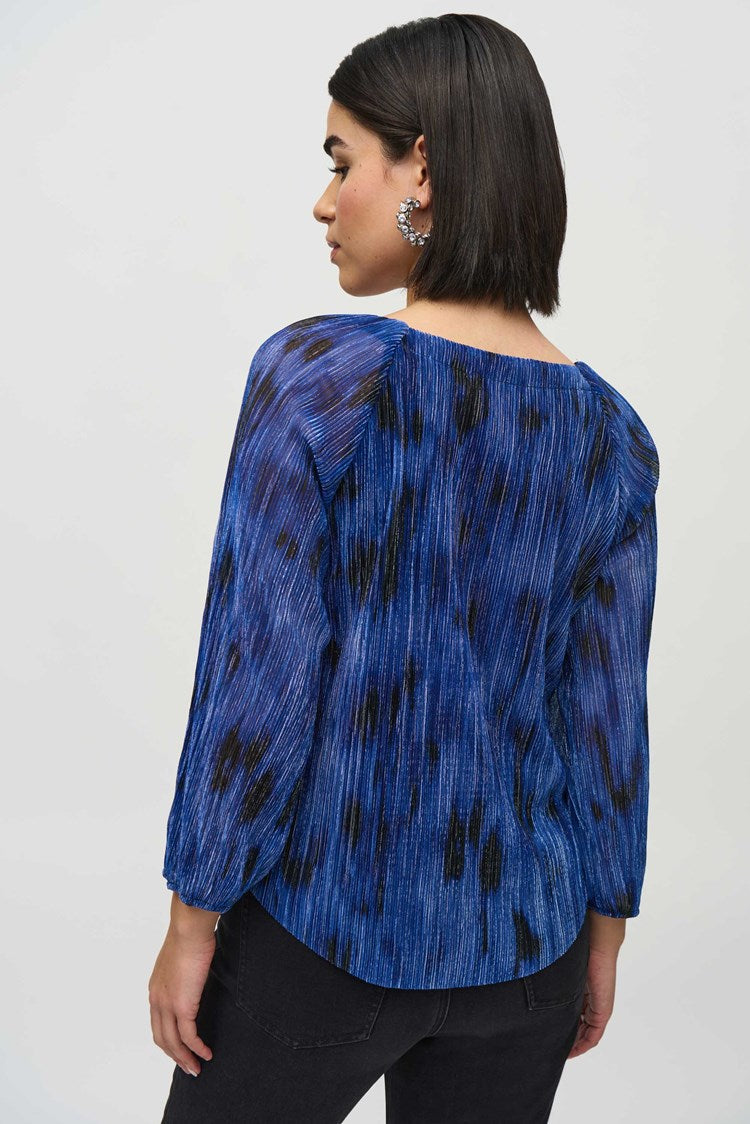 A person with shoulder-length black hair is standing against a plain background, wearing the Joseph Ribkoff Pleated Knit Abstract Print Flared Top 244105, which is blue with long sleeves and features a textured, pleated design. They are also wearing dark pants and are looking slightly to the side with a calm expression.