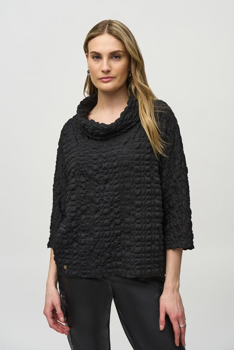 A woman stands against a plain white background, wearing a black Woven Jacquard Boxy Top 244069 by Joseph Ribkoff with three-quarter sleeves, black leather pants with side pockets, and black heeled ankle boots. Her left hand is on her hip and her right arm hangs by her side. She has long, wavy hair.