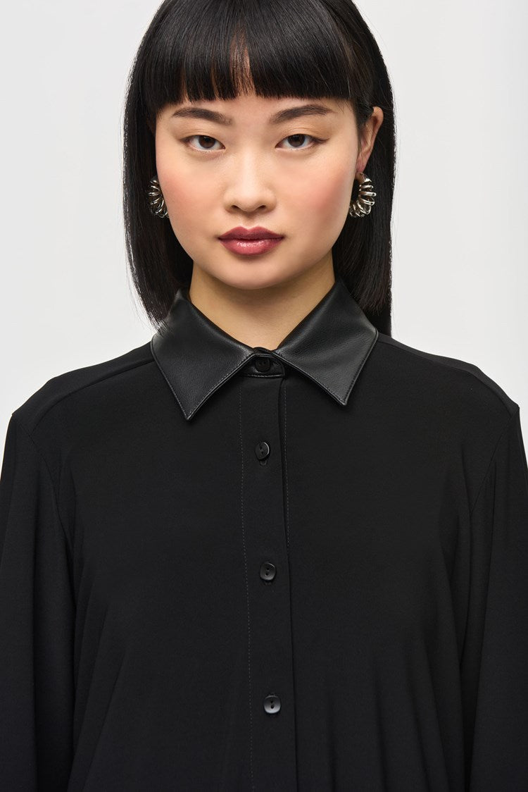 A person with long black hair and bangs poses against a plain background, wearing the Silky Knit Fit and Flare Blouse 243204 by Joseph Ribkoff. The blouse features a leatherette faux collar and leather cuffs, paired elegantly with black pants. Their expression is neutral, and one hand rests gently on their opposite arm, just above the elbow.