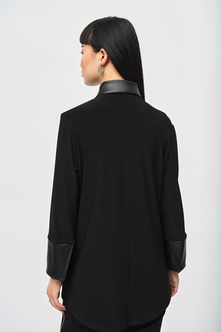 A person with long black hair and bangs poses against a plain background, wearing the Silky Knit Fit and Flare Blouse 243204 by Joseph Ribkoff. The blouse features a leatherette faux collar and leather cuffs, paired elegantly with black pants. Their expression is neutral, and one hand rests gently on their opposite arm, just above the elbow.