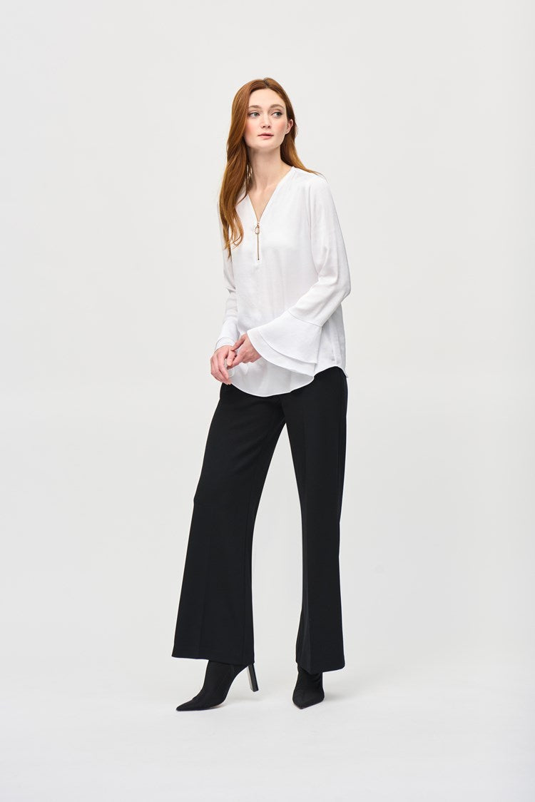 A woman with long reddish-brown hair wearing the Joseph Ribkoff Satin Boxy Top With Ruffled Sleeve 243285, which features a V-shaped neckline and long ruffled sleeves, stands against a plain white background. She is looking directly at the camera with a neutral expression, one hand by her side and the other resting on her hip.
