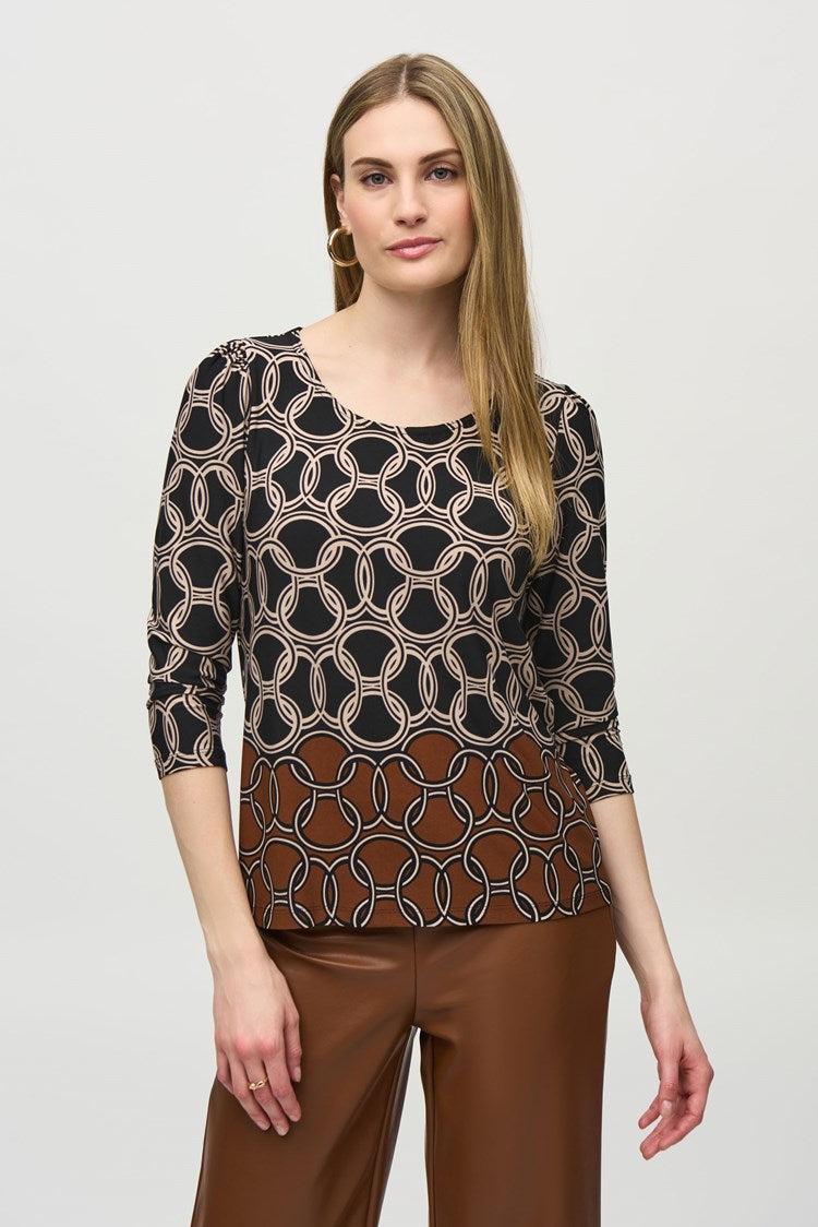 A woman stands against a plain white background. She is wearing the Silky Knit Chain Border Print Top by Joseph Ribkoff, which has three-quarter sleeves and an abstract white pattern. She pairs it with brown wide-leg leather pants and black ankle boots. She is looking straight at the camera, with her arms relaxed by her sides.