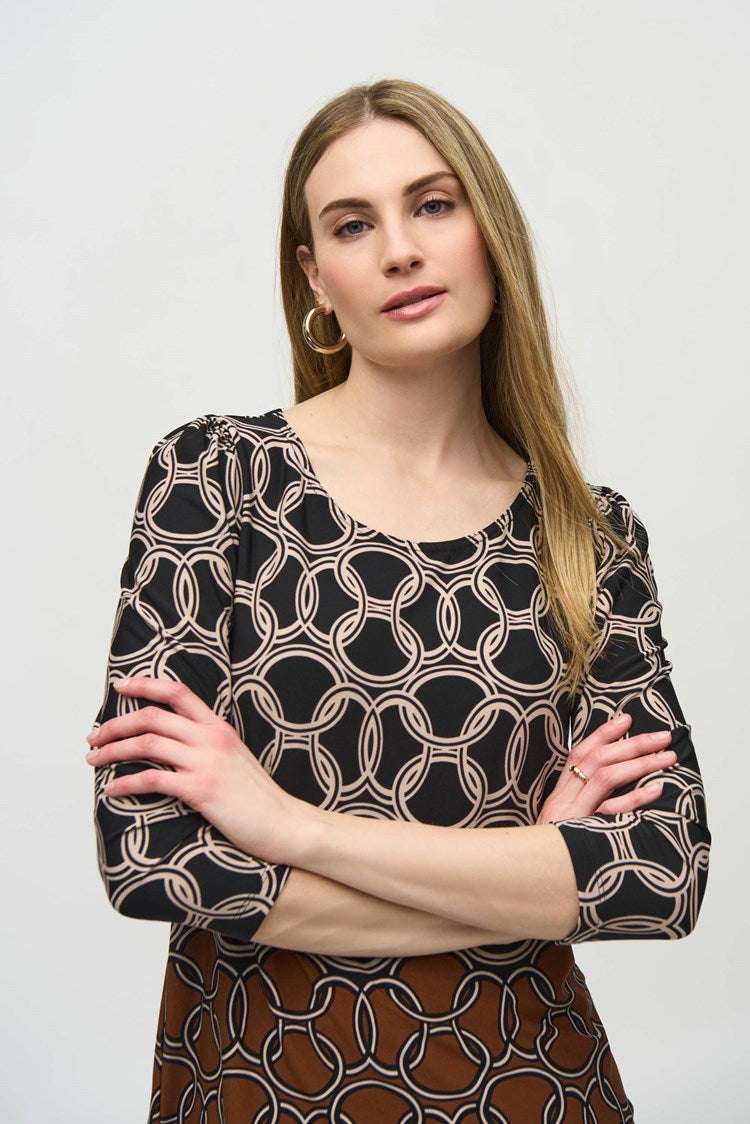 A woman stands against a plain white background. She is wearing the Silky Knit Chain Border Print Top by Joseph Ribkoff, which has three-quarter sleeves and an abstract white pattern. She pairs it with brown wide-leg leather pants and black ankle boots. She is looking straight at the camera, with her arms relaxed by her sides.