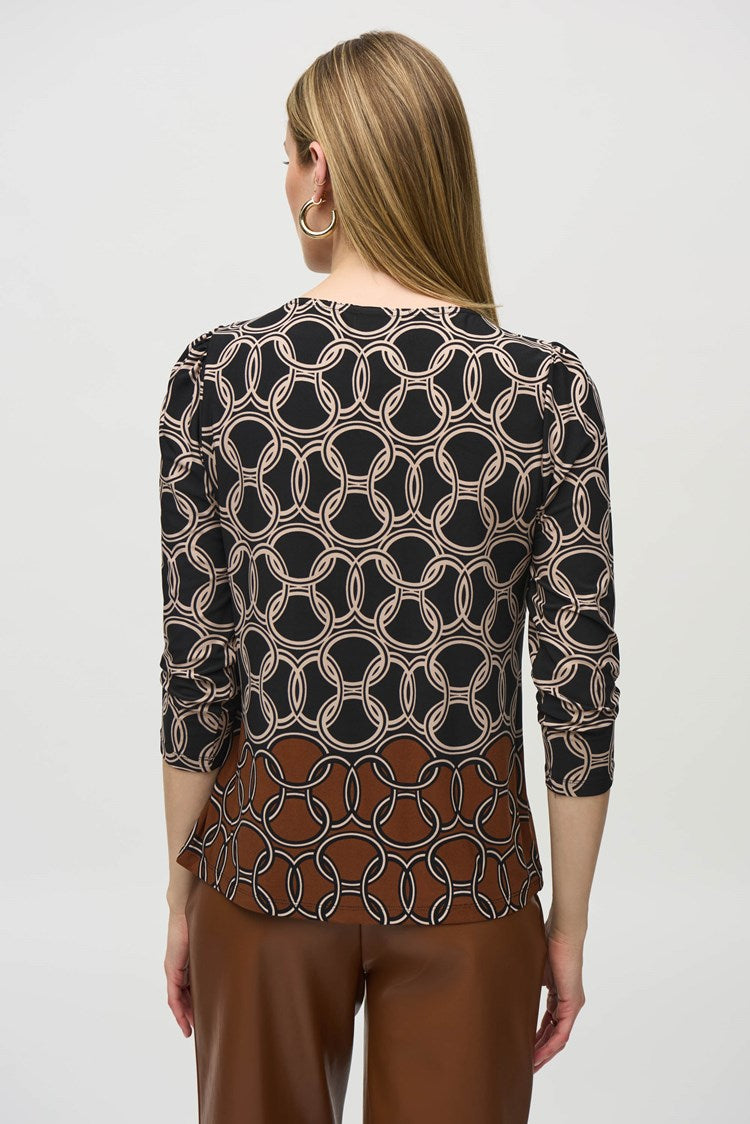A woman stands against a plain white background. She is wearing the Silky Knit Chain Border Print Top by Joseph Ribkoff, which has three-quarter sleeves and an abstract white pattern. She pairs it with brown wide-leg leather pants and black ankle boots. She is looking straight at the camera, with her arms relaxed by her sides.