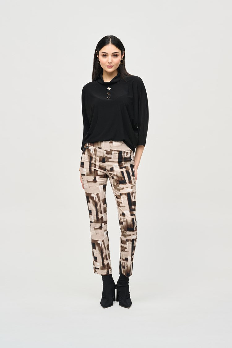 A woman with long dark hair stands against a plain background. She is wearing a black top with buttons at the neckline and Joseph Ribkoff's Millennium Abstract Print Slim Pull-On Pants, featuring beige, brown, and black geometric designs. Her slim silhouette is enhanced by black ankle boots, and her left hand rests casually in her pocket.