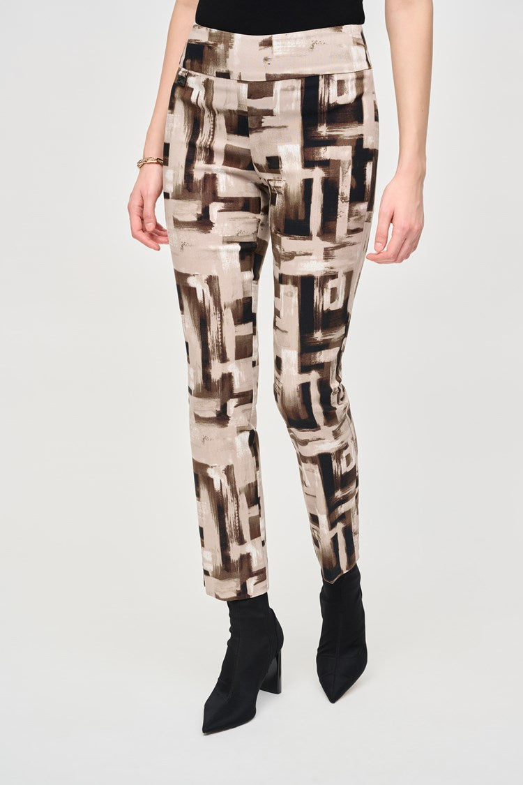 A woman with long dark hair stands against a plain background. She is wearing a black top with buttons at the neckline and Joseph Ribkoff's Millennium Abstract Print Slim Pull-On Pants, featuring beige, brown, and black geometric designs. Her slim silhouette is enhanced by black ankle boots, and her left hand rests casually in her pocket.