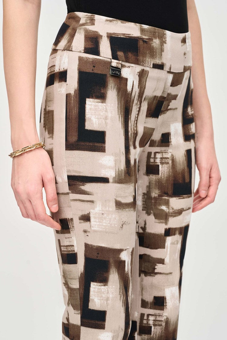 A woman with long dark hair stands against a plain background. She is wearing a black top with buttons at the neckline and Joseph Ribkoff's Millennium Abstract Print Slim Pull-On Pants, featuring beige, brown, and black geometric designs. Her slim silhouette is enhanced by black ankle boots, and her left hand rests casually in her pocket.