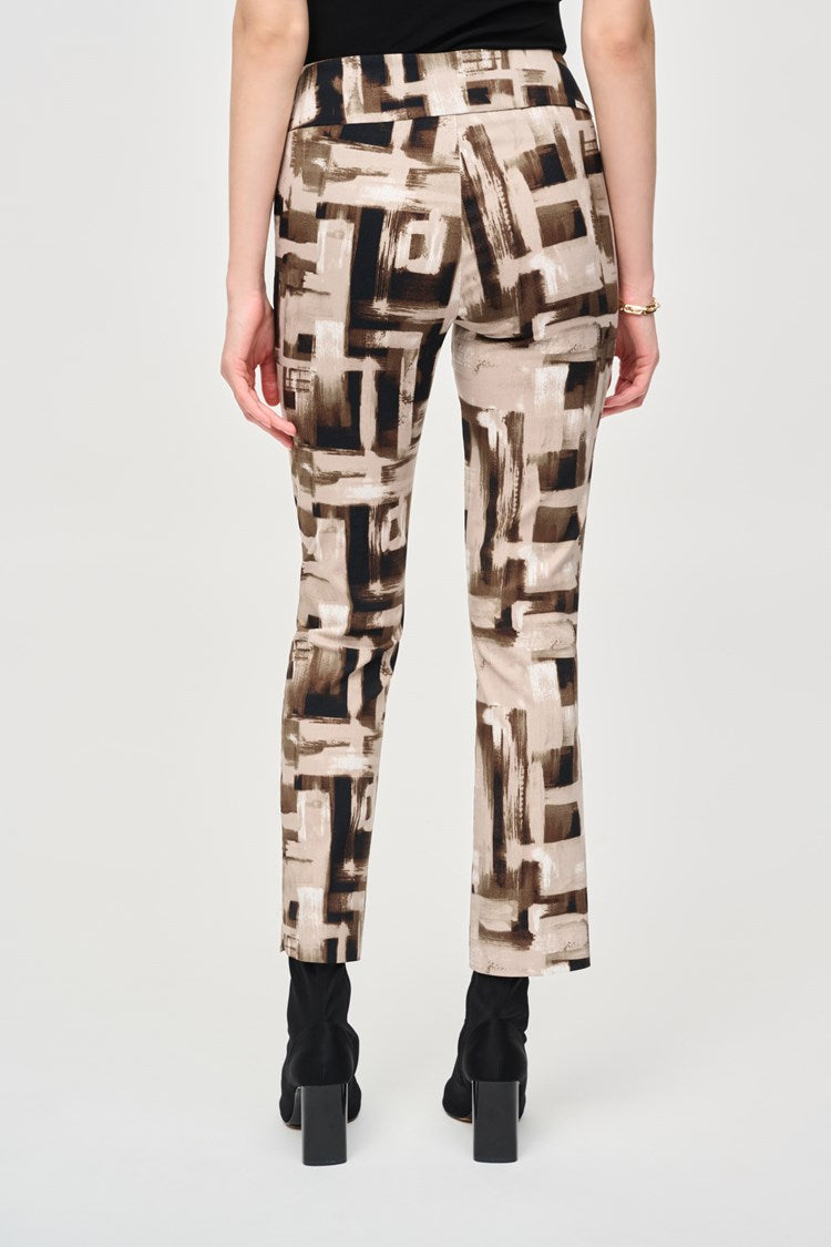 A woman with long dark hair stands against a plain background. She is wearing a black top with buttons at the neckline and Joseph Ribkoff's Millennium Abstract Print Slim Pull-On Pants, featuring beige, brown, and black geometric designs. Her slim silhouette is enhanced by black ankle boots, and her left hand rests casually in her pocket.
