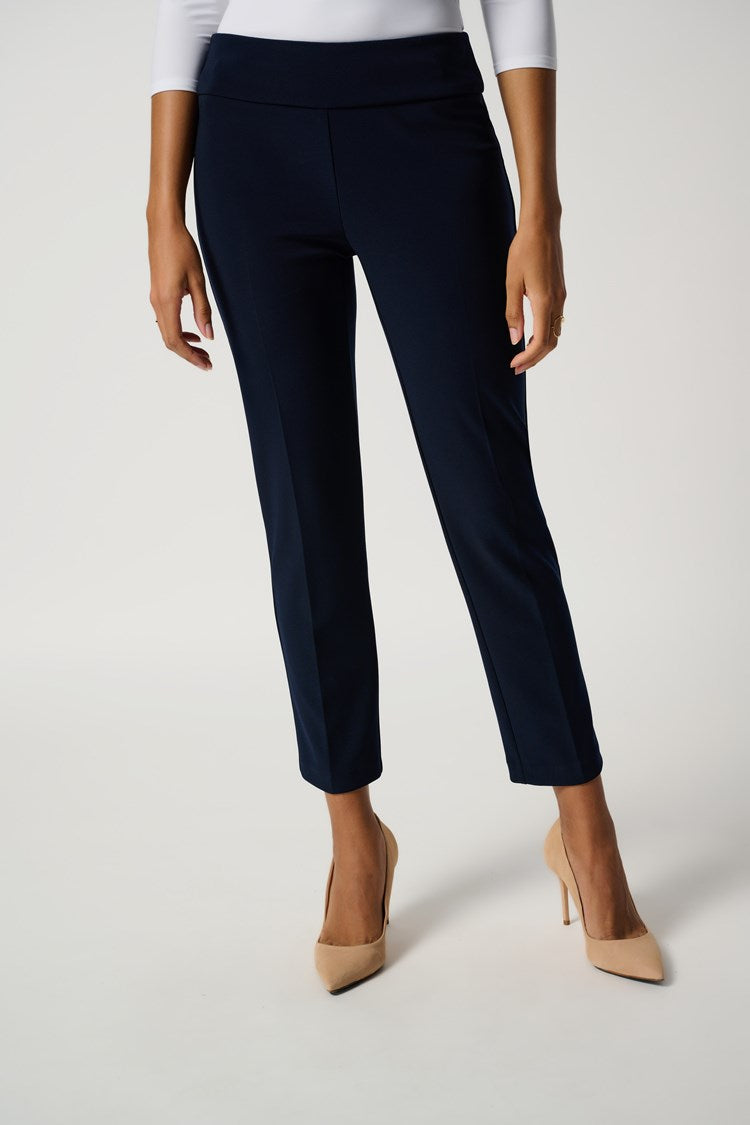A woman with dark curly hair is standing against a plain white background. She is wearing a white, three-quarter sleeve top made of silky knit fabric, the "Classic Cropped Pant - Core Colors" by Joseph Ribkoff in dark blue, and beige high heels. Her arms are crossed casually, and she has a neutral expression.