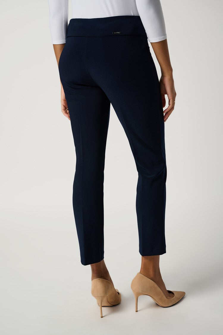 A woman with dark curly hair is standing against a plain white background. She is wearing a white, three-quarter sleeve top made of silky knit fabric, the "Classic Cropped Pant - Core Colors" by Joseph Ribkoff in dark blue, and beige high heels. Her arms are crossed casually, and she has a neutral expression.
