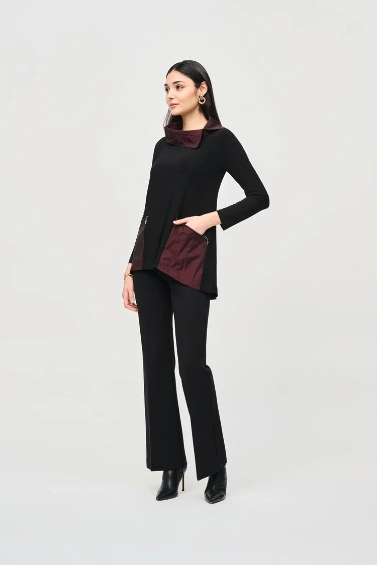 A woman with long dark hair stands confidently in a Joseph Ribkoff black outfit featuring a maroon collar and maroon panels at the waist. She wears the Silky Knit And Jacquard Memory Tunic paired with black pants and ankle boots. The background is plain light gray.