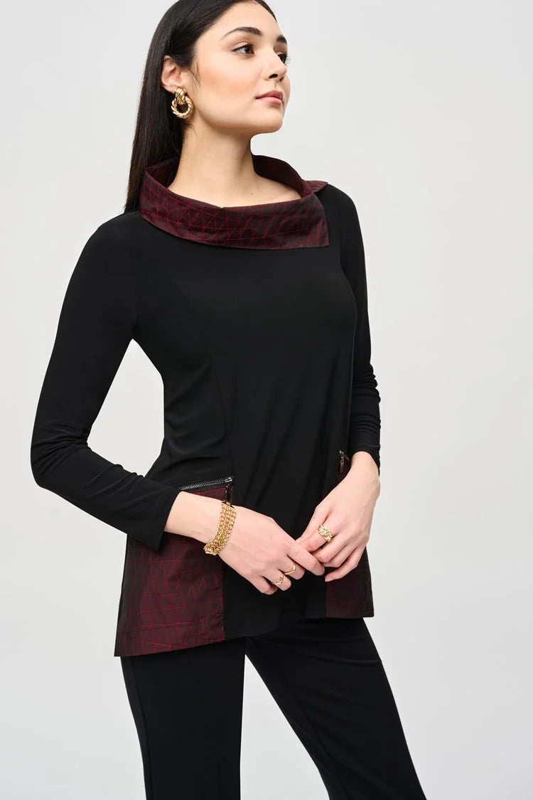 A woman with long dark hair stands confidently in a Joseph Ribkoff black outfit featuring a maroon collar and maroon panels at the waist. She wears the Silky Knit And Jacquard Memory Tunic paired with black pants and ankle boots. The background is plain light gray.