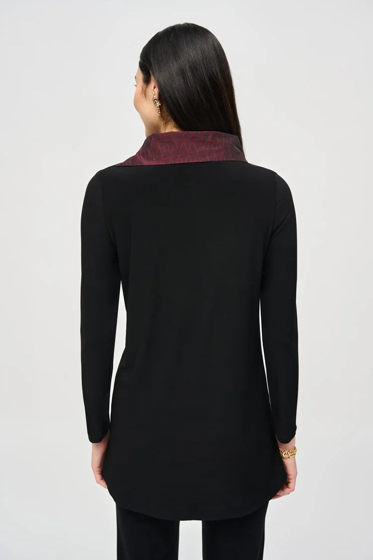 A woman with long dark hair stands confidently in a Joseph Ribkoff black outfit featuring a maroon collar and maroon panels at the waist. She wears the Silky Knit And Jacquard Memory Tunic paired with black pants and ankle boots. The background is plain light gray.