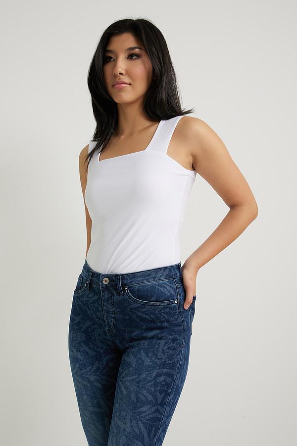 A woman with long dark hair stands against a plain background, smiling. She is wearing a white sleeveless top, Joseph Ribkoff's Leaf Print Classic Slim Jeans - 222930, and beige high heels. Her arms are relaxed by her sides.