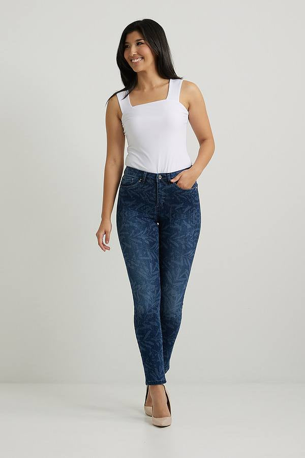 A woman with long dark hair stands against a plain background, smiling. She is wearing a white sleeveless top, Joseph Ribkoff's Leaf Print Classic Slim Jeans - 222930, and beige high heels. Her arms are relaxed by her sides.