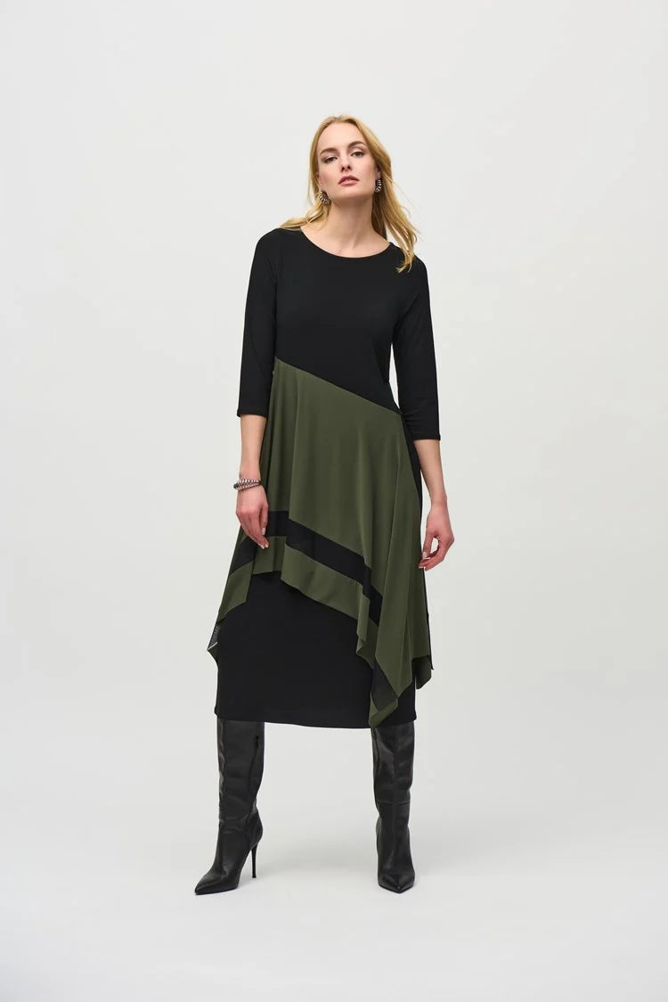A woman stands against a plain background, wearing the Joseph Ribkoff Silky Knit Colour Block Handkerchief Dress 244186 in black and olive green. The asymmetrical dress, made from a silky knit fabric, features three-quarter sleeves and a handkerchief hemline that ends at mid-calf length. She completes her outfit with black knee-high boots. Her expression is neutral, and her hands rest at her sides.