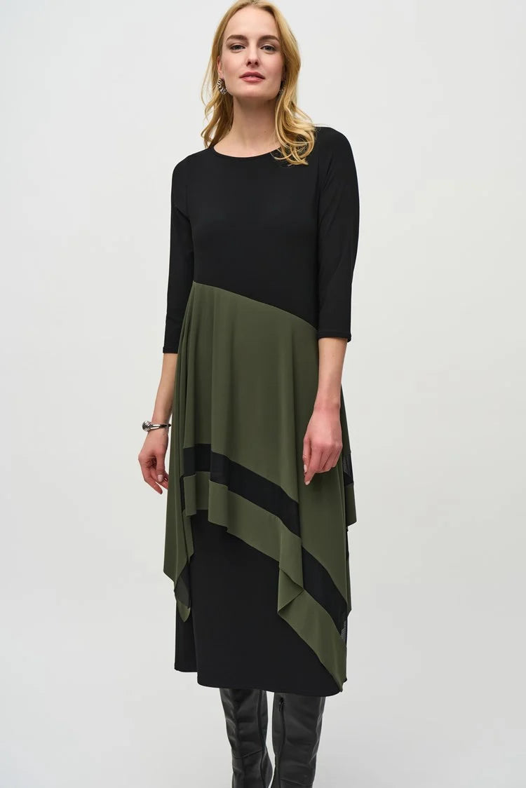 A woman stands against a plain background, wearing the Joseph Ribkoff Silky Knit Colour Block Handkerchief Dress 244186 in black and olive green. The asymmetrical dress, made from a silky knit fabric, features three-quarter sleeves and a handkerchief hemline that ends at mid-calf length. She completes her outfit with black knee-high boots. Her expression is neutral, and her hands rest at her sides.
