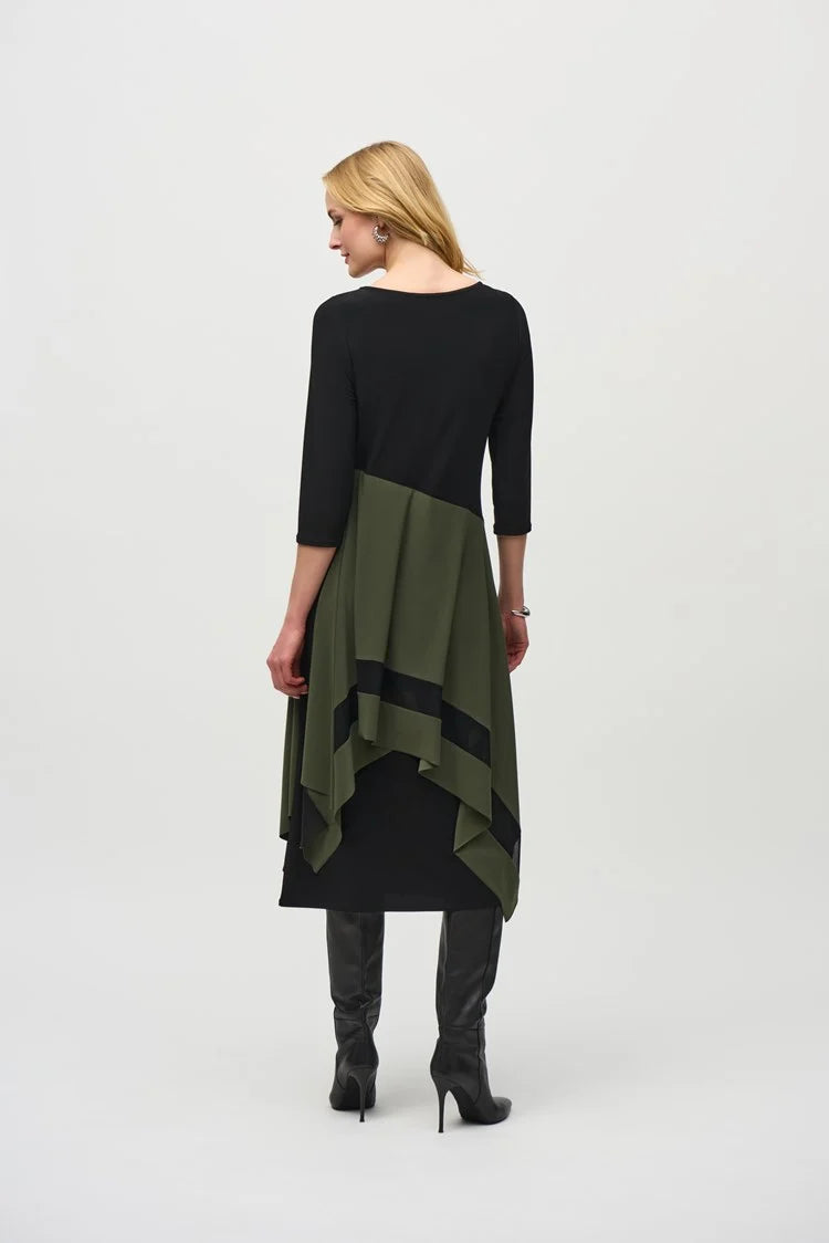 A woman stands against a plain background, wearing the Joseph Ribkoff Silky Knit Colour Block Handkerchief Dress 244186 in black and olive green. The asymmetrical dress, made from a silky knit fabric, features three-quarter sleeves and a handkerchief hemline that ends at mid-calf length. She completes her outfit with black knee-high boots. Her expression is neutral, and her hands rest at her sides.