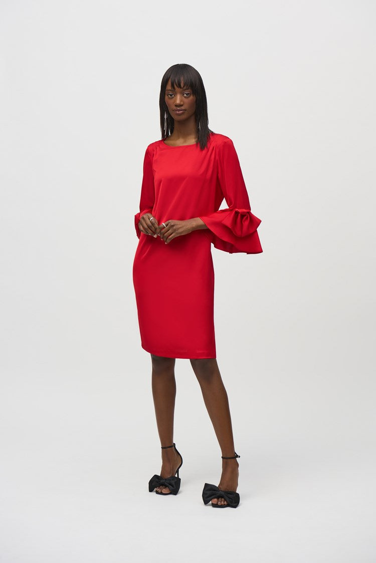 A woman stands against a plain white background, wearing the Joseph Ribkoff Satin Ruffle Sleeve Straight Dress (style number 244235) in bright red. This heavy satin dress features an elegant boat neckline, long sleeves with ruffle details at the cuffs, and a back zipper opening. She has shoulder-length dark hair and wears black open-toed heels with bow accents, posing with her hands gently clasped.
