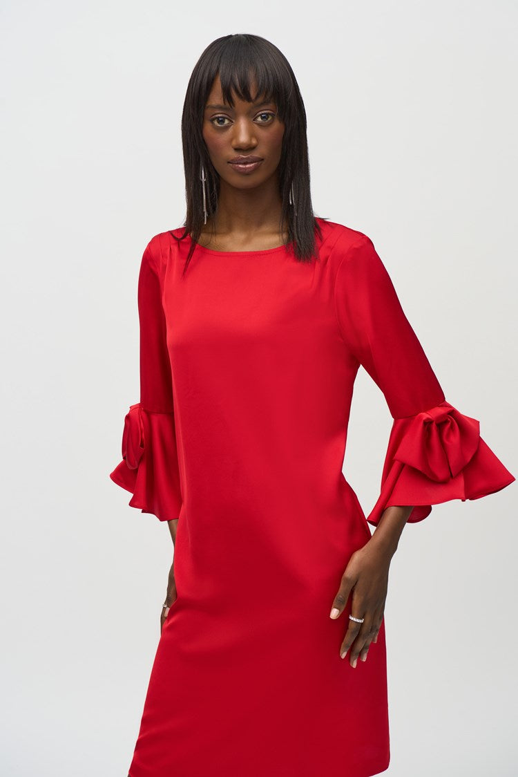 A woman stands against a plain white background, wearing the Joseph Ribkoff Satin Ruffle Sleeve Straight Dress (style number 244235) in bright red. This heavy satin dress features an elegant boat neckline, long sleeves with ruffle details at the cuffs, and a back zipper opening. She has shoulder-length dark hair and wears black open-toed heels with bow accents, posing with her hands gently clasped.