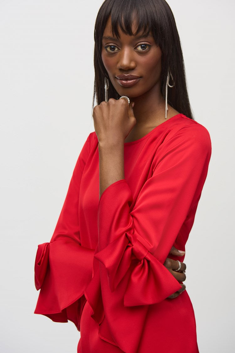 A woman stands against a plain white background, wearing the Joseph Ribkoff Satin Ruffle Sleeve Straight Dress (style number 244235) in bright red. This heavy satin dress features an elegant boat neckline, long sleeves with ruffle details at the cuffs, and a back zipper opening. She has shoulder-length dark hair and wears black open-toed heels with bow accents, posing with her hands gently clasped.