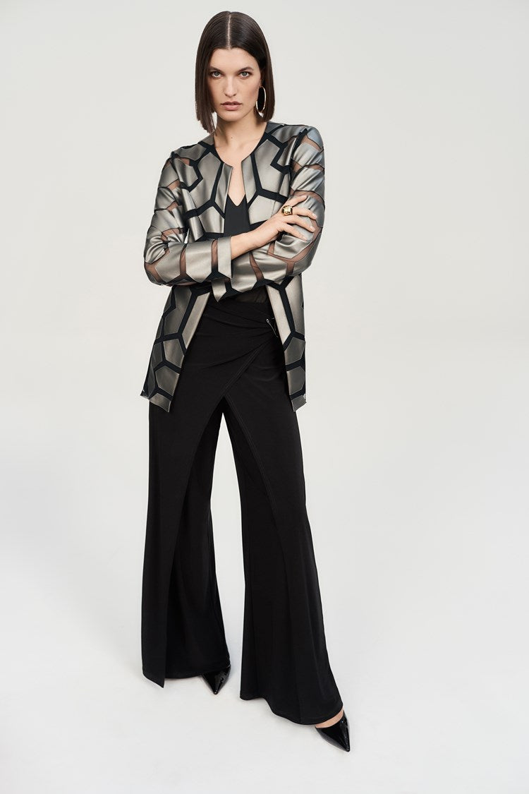 A woman stands confidently against a plain background. She is wearing a long-sleeved, geometric-patterned jacket over a black top and Joseph Ribkoff’s Silky Knit Wide-Leg Wrap Pants with slits. She completes her look with black pointed-toe heels, large hoop earrings, and an eye-catching asymmetrical wrap design.