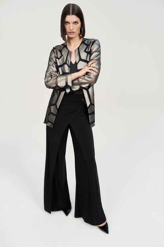 A woman stands confidently against a plain background. She is wearing a long-sleeved, geometric-patterned jacket over a black top and Joseph Ribkoff’s Silky Knit Wide-Leg Wrap Pants with slits. She completes her look with black pointed-toe heels, large hoop earrings, and an eye-catching asymmetrical wrap design.