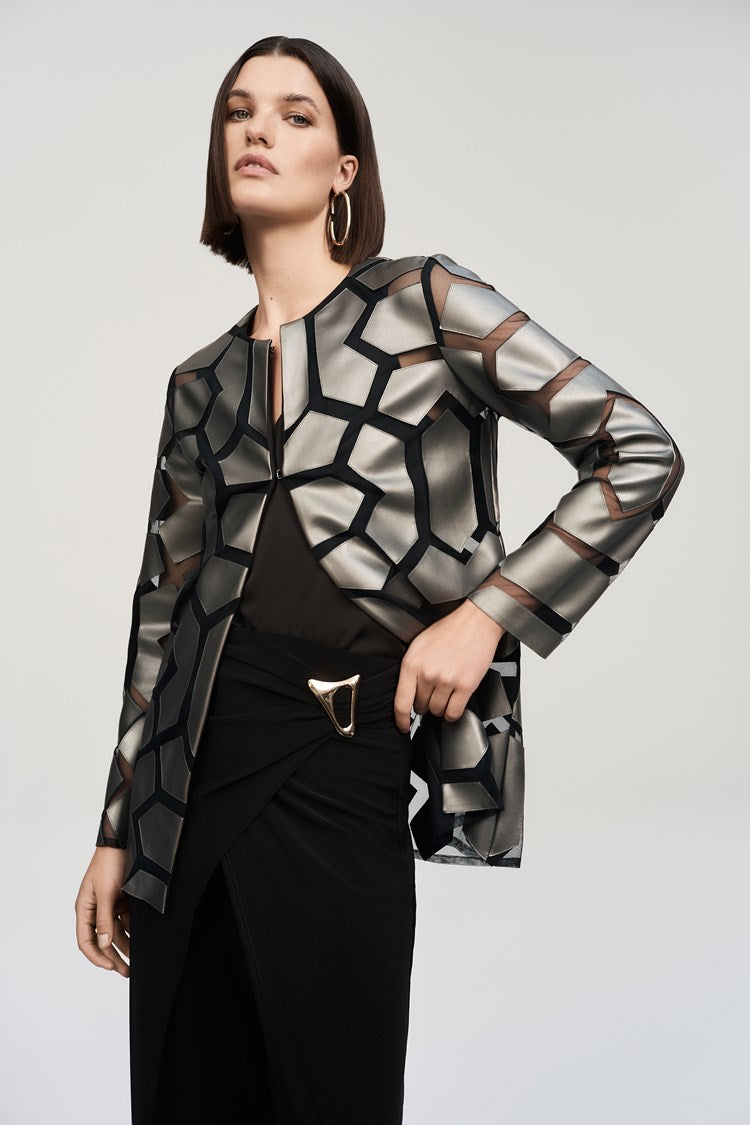 A woman stands confidently against a plain background. She is wearing a long-sleeved, geometric-patterned jacket over a black top and Joseph Ribkoff’s Silky Knit Wide-Leg Wrap Pants with slits. She completes her look with black pointed-toe heels, large hoop earrings, and an eye-catching asymmetrical wrap design.